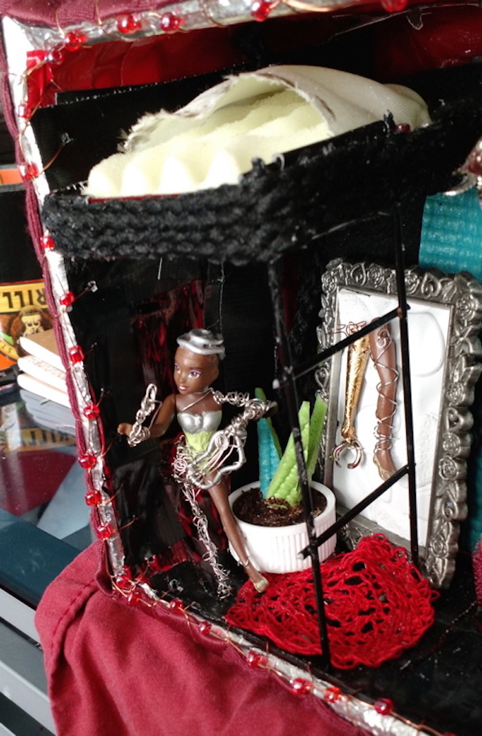 Photo of the sample mockup of Sessaris's apartment, removed from the shelving, seen from the open side. Sessaris, standing under the bunk bed, is made from a brown-skinned miniature princess doll, with her prostheses made of steel wire. A plant made of green yoga-mat foam grows behind her, planted in a plastic bottle cap filled with a dried mix of glue and ground coffee. A tiny red rug that I crocheted from embroidery floss is beneath her feet. In this version, a realistic prosthetic leg made from the doll's own leg, and a prosthetic arm with a robotic claw made from a jeweler's lobster clasp, hang in a miniature picture frame that stands behind her.