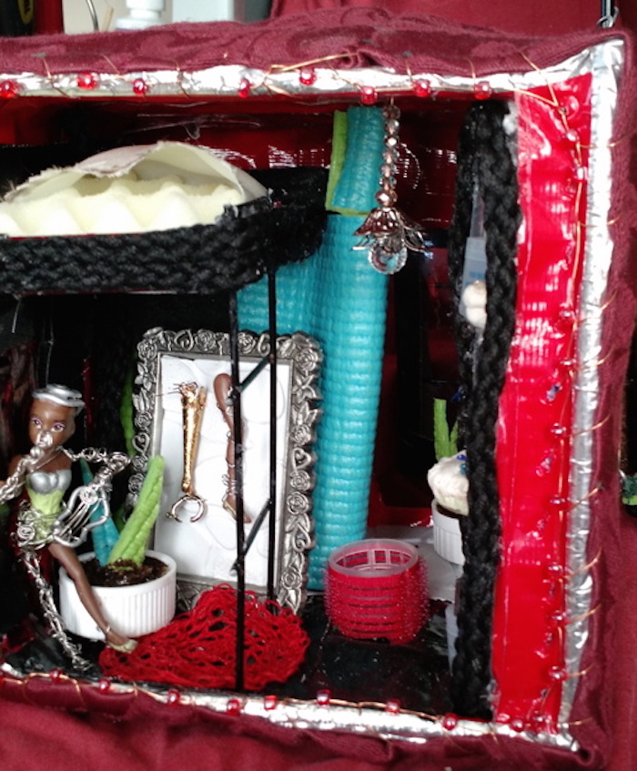 Photo of the sample mockup of Sessaris's apartment, removed from the shelving, seen from the open side. This angle shows more of the center of the room, revealing a light fixture made of beads, and a large red hair roller protruding from the floor, which, in this version, forms the tube that leads into the downstairs half of the apartment.