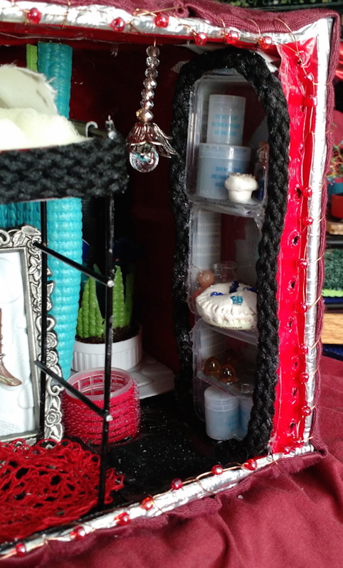 Photo of the sample mockup of Sessaris's apartment, removed from the shelving, seen from the open side. This view focuses on a shelf of food, including several cans made from desiccant canisters, a muffin and a bottle-cap pie made with clay and beads, and a pill-blister carton of clear yellow eggs.