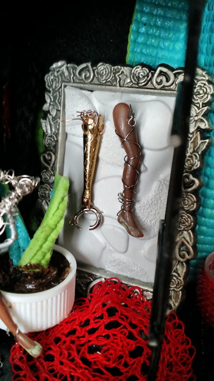 A close-up view of the spare prostheses that are visible behind Sessaris. A realistic prosthetic leg made from the doll's own leg, wrapped in steel wire, and a prosthetic arm with a robotic claw made from a jeweler's lobster clasp, hang in a miniature picture frame. The picture frame is pewter-colored and decorated with images of hearts and roses.