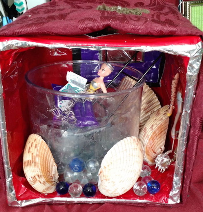 Photo of the sample mockup of Kit's apartment, removed from the shelving, seen from the open side. Room is dominated by a large cylindrical glass container, filled with marbles and shells, and topped with clear plastic to suggest water. Decorative seashells flank the outside of the container, and spare prostheses hang on the wall near it. A ladder from the ceiling leads down into it. A window is visible behind the ladder. Kit is made from a plastic doll like Sessaris, this one light-skinned, missing both legs, and having both arms. In this version her mermaid tail is made entirely of steel wire, and she is floating on a purple raft instead of swimming. She holds a palette and a book of paper in which she is painting.