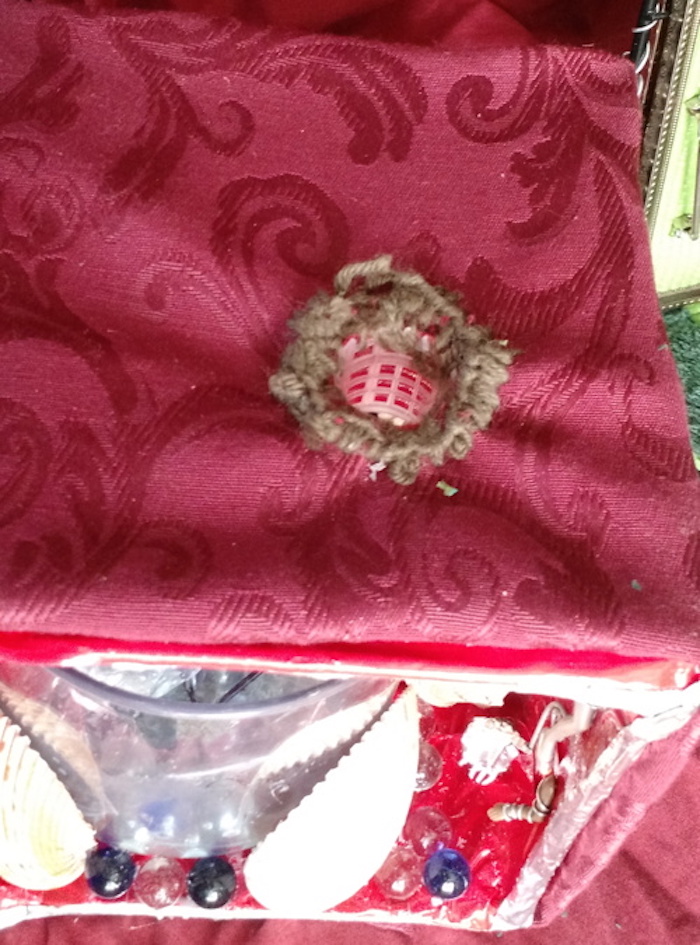 Photo of the sample mockup of Kit's apartment, removed from the shelving, seen from the top. This view shows that the roof is made of brocaded red fabric, and the opening to the tube is hemmed in brown embroidery thread.