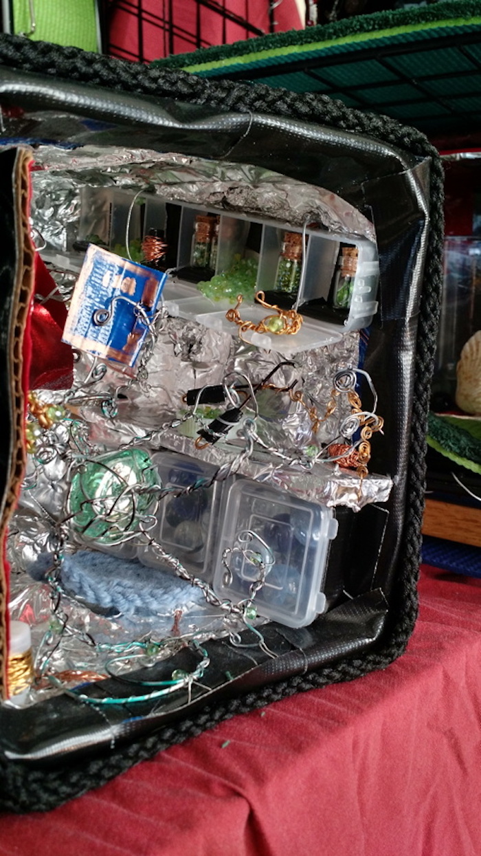 Photo of the sample mockup of the apartment, removed from the shelving, seen from the open side. This view focuses on the workbench. The cubbies hold mostly tiny corked glass bottles, filled with transparent green beads. You can also see in this view that the front of the cube is outlined in a frame of black rope, and there is a light blue crocheted rug on the floor.