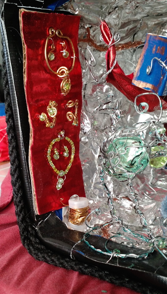 Close-up of the stand with jewelry hanging just inside the cube, on the left wall. It is made of red satin and covered with brass jewelry set with green stones. At the top, a collar necklace with a stone in the center of a spiralling pendant, and a matching pair of spiralling earrings. Below that, two cuff bracelets of thick brass wire with much thinner wire wrapped around them; the nearer one has green stones on both ends. At the bottom, a necklace strung with many tiny green beads, with a central pendant made of three faceted green crystal beads one above the next, and two matching earrings, each one a short string of tiny beads ending in a single faceted crystal bead. Nearby on the floor is another spool of brass wire.