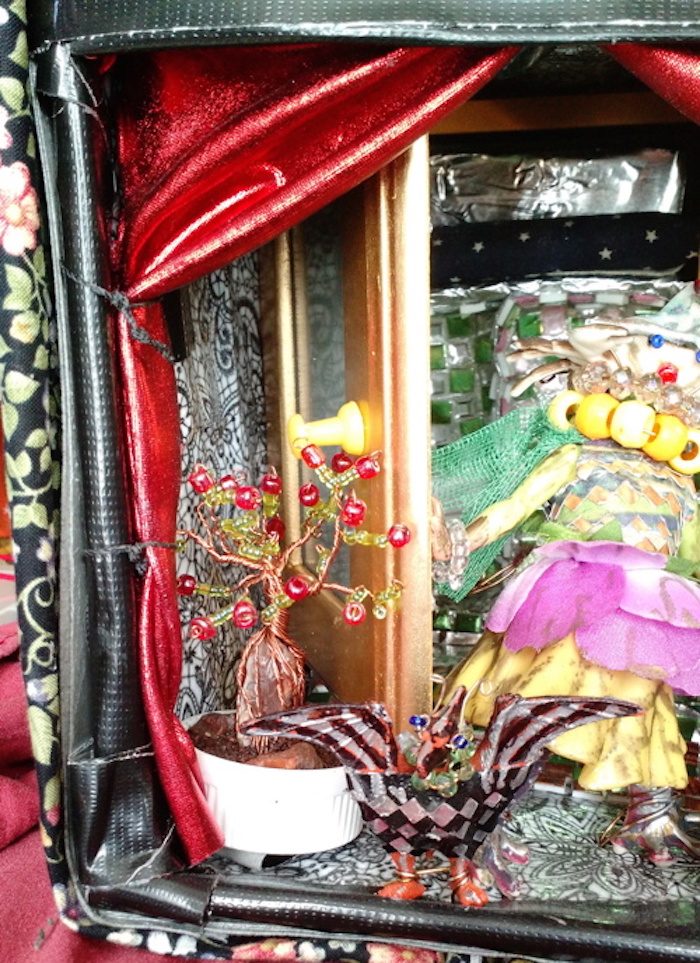 Photo of the sample mockup of the apartment, removed from the shelving, seen from the open side. This view focuses on Singer the bird, and the leftmost potted tree. Singer has eyes made from blue beads, and wears a necklace made of green faceted glass beads. The tree has branches made from twisted copper wire, with the leaves and berries made from green and red glass beads; the roots wrap around a large rough agate stone that is half-submerged in the coffee soil.