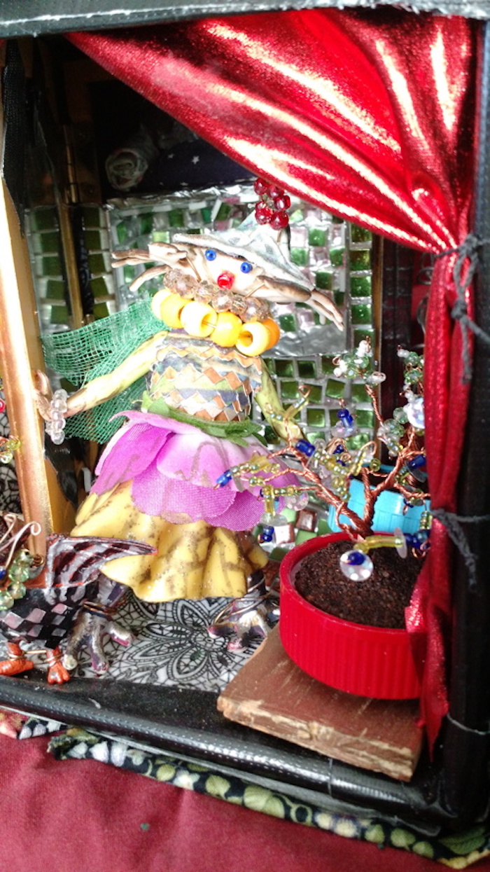 Photo of the sample mockup of the apartment, removed from the shelving, seen from the open side. This view focuses the rightmost potted tree. It is also made of copper wire and potted in coffee grounds in a red bottle cap. It has leaves and berries made from green and blue beads, as well as flowers made of silver sequins. Around it you can also see that the open curtain is made of red satin and tied against the wall with black thread.